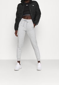 Zwarte pufferjas met een hoge kraag, grijze trainingsbroek met boorden en witte sneakers. De outfit bestaat uit een cropped zwart topje met logo-accenten.