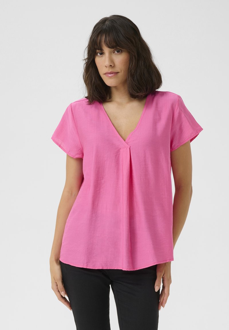Roze blouse met korte mouwen, een V-hals en plooidetail, gemaakt van een lichte, gestructureerde stof. Draagt met donkere broek.