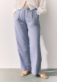 Hele sinised linased laiad püksid esikokkupandega ja nööpkinnitusega. Kandmisel on pruunid punutud sandaalid. Minimalistlik disain, sile tekstuur.