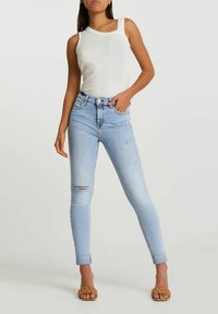 Top de tirantes blanco acanalado combinado con jeans skinny de tiro alto en azul claro, con una rodilla desgastada y dobladillo acortado, complementado con sandalias marrones.