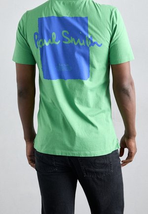 Personne portant un T-shirt vert avec un grand carré bleu et le texte "Paul Smith" au dos, assorti à un jean sombre.
