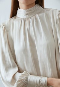 Lys beige bluse med høy, samlet halslinning og voluminøse pufferm. Stoffet er glatt og glansfullt, som fremhever myke folder og subtil glans.