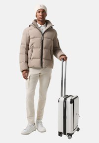 Mann in beiger Daunenjacke, weißen Cargo-Hosen und Strickmütze, der mit einem weißen Rollkoffer vor einem weißen Hintergrund steht.