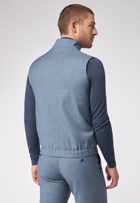 Hellblaues, strukturiertes Weste mit hohem Kragen, kombiniert mit einem dunkelblauen Langarmhemd. Verfügt über einen elastischen Taillenbund und ein figurbetontes Design.