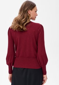 Kobieta z falującymi brązowymi włosami, nosząca złote kolczyki w kształcie kół oraz bordowy sweter z długim rękawem i bufkami, w czarnej spódnicy, odwrócona plecami.