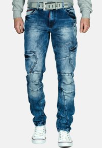 Pantaloni cargo in denim blu chiaro con lavaggio sfumato, multiple tasche e dettagli di cucitura distintivi; abbinati a sneaker bianche.