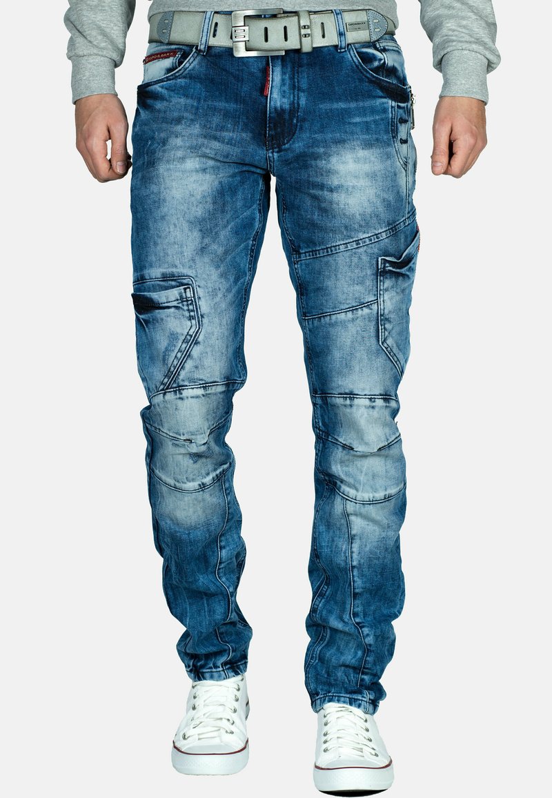 Pantaloni cargo in denim blu chiaro con lavaggio sfumato, multiple tasche e dettagli di cucitura distintivi; abbinati a sneaker bianche.