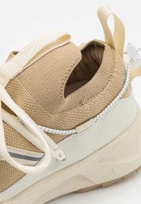 Beżowe tenisówki z teksturowaną cholewką, elastycznymi sznurówkami i białą teksturowaną podeszwą. Posiadają pętelkę na pięcie oraz kontrastowe szwy.