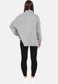 Vrouw staand met de rug naar de kijker, gekleed in een losse grijze gebreide trui, zwarte leggings en witte sneakers tegen een eenvoudige achtergrond.