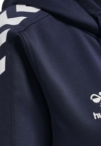 Felpa blu navy realizzata in morbido tessuto sintetico, con un logo bianco e un ampio motivo a chevron sulla spalla. Cappuccio con coulisse regolabile.