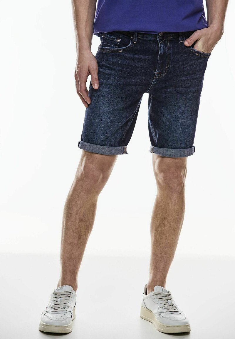 Džíny šortky v tmavě modré barvě s ohrnutými nohavicemi, s klasickým pětiokrovým designem a jemnými detaily prošívání. Nosené s bílými teniskami.