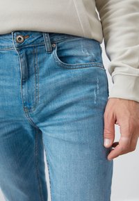 Lyseblå denimjeans med en let slidt finish, der har orange syninger, en enkelt frontlomme og en knaplukning i taljen.