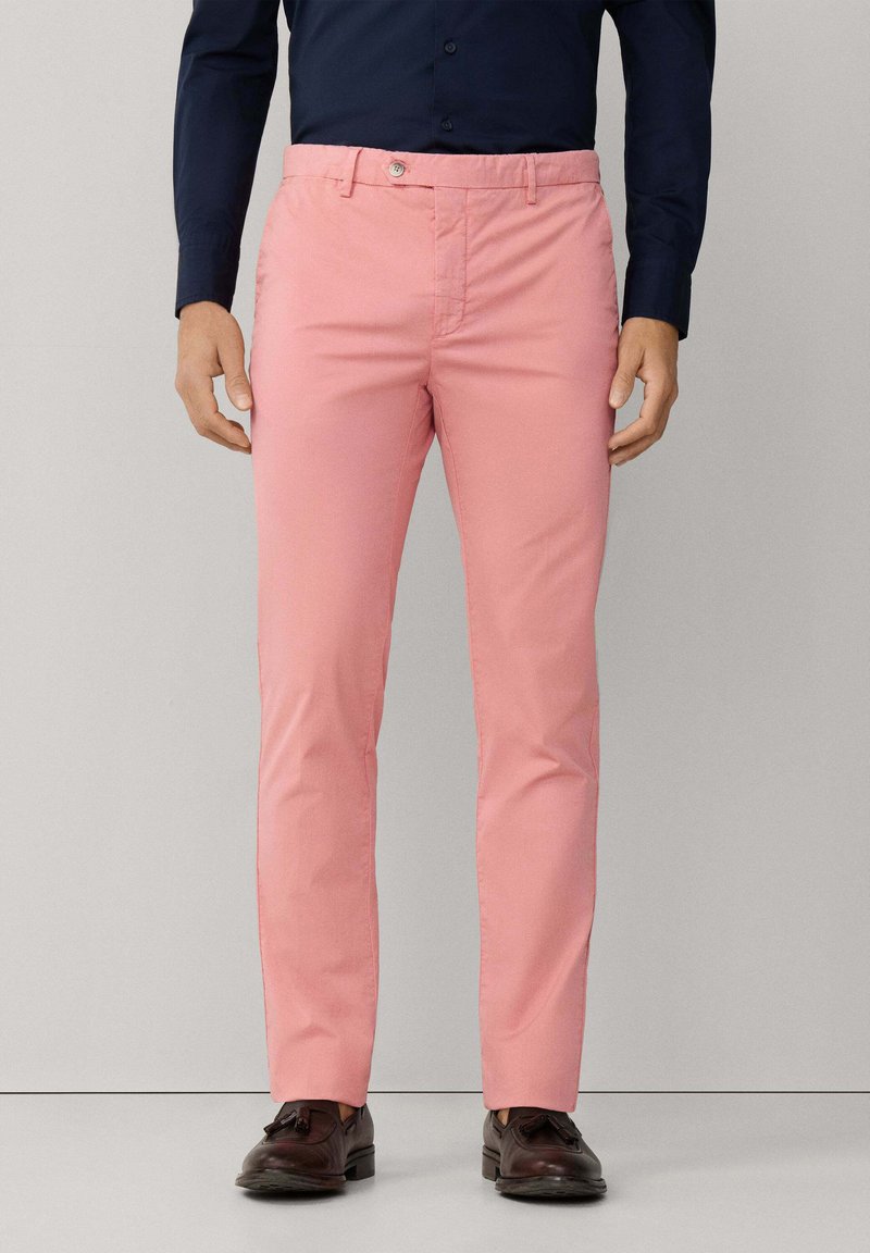 Uomo che indossa pantaloni rosa slim-fit, camicia blu scuro con bottoni e mocassini scuri con nappine, in piedi davanti a uno sfondo semplice.