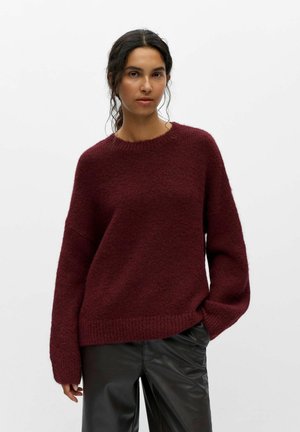 Femme aux cheveux bouclés foncés portant un pull ample bordeaux et un pantalon en cuir noir, debout sur un fond clair uni.