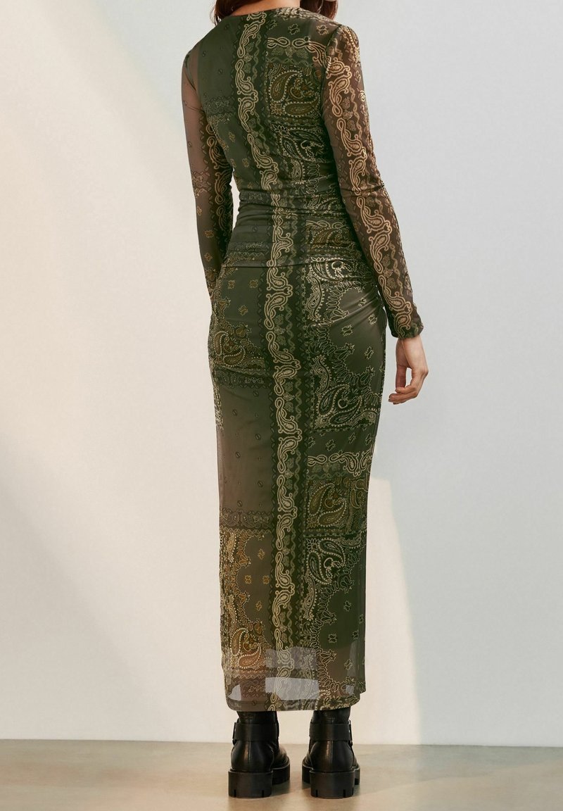 Femme portant une longue robe transparente à motifs vert olive avec des dessins cachemire beige, associée à des bottines noires épaisses, debout contre un mur uni.