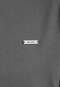 Grau T-Shirt-Stoff mit einer glatten Textur, an der Vorderseite mit einem kleinen silbernen Etikett mit der Gravur "SMILODOX" versehen.