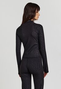 Donna di spalle che indossa una maglietta a maniche lunghe nera e pantaloni con un sottile motivo leopardato scuro su uno sfondo chiaro e semplice.