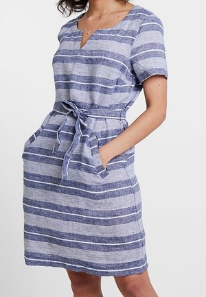 Frau trägt ein kurzärmeliges, knielanges blau-grau gestreiftes Kleid mit Taillenband und Taschen, steht mit den Händen in den Taschen.