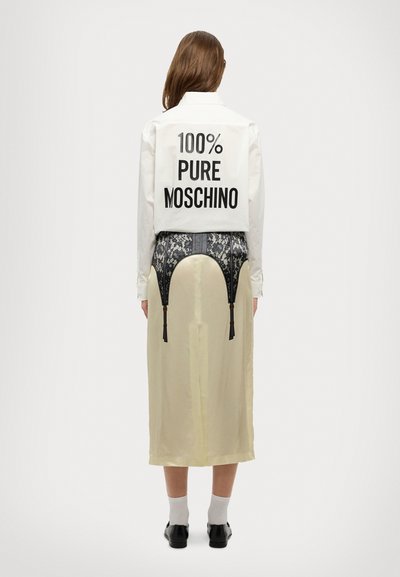 Cămașă albă cu textul "100% PURE MOSCHINO" pe spate, combinată cu o fustă din satin bej, cu dantelă neagră și bretele. Pantofi negri, cu talpă plată.