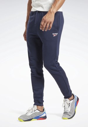 Mand iført marineblå Reebok joggingbukser og hvide sportssko med flerfarvede såler, stående mod en ensfarvet hvid baggrund.