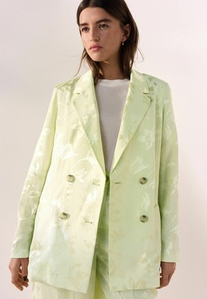 Young woman wearing a light green floral double-breasted blazer over a white top, gazing slightly to the side against a plain background.