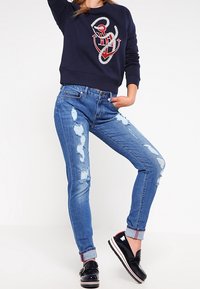 Woman wearing distressed blue jeans, navy sweatshirt with red and white anchor design, and black platform loafers, posing with one leg bent.