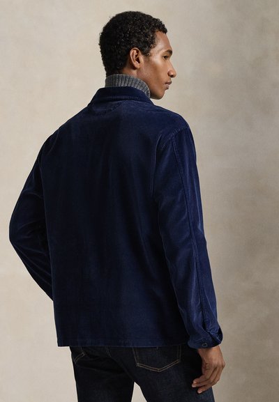 Άνδρας, φανερός από πίσω, φοράει σκούρο μπλε σακάκι από κορντόρι πάνω από γκρι πουλόβερ με ζιβάγκο και μαύρο τζιν.