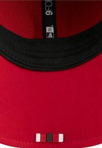 Rote Kappe mit schwarzer Unterseite; verfügt über einen strukturierten Stoff. Betont durch ein kleines Logotag und drei Farbblöcke (rot, schwarz, weiß) am Rand.