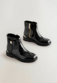 Schwarze Lackleder-Stiefeletten mit seitlichem Reißverschluss, einem strukturierten schwarzen Obermaterial und einer dekorativen Schleifenverzierung an der Vorderseite. Mit goldenen Akzenten.