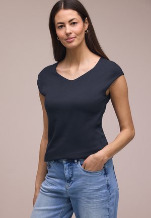 Mujer con cabello largo y castaño que lleva pendientes de aro dorados, un top negro sin mangas y jeans azul claro, de pie con una mano en el bolsillo.