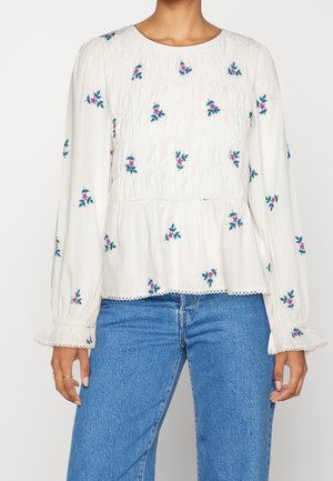 Vrouw draagt een witte blouse met lange mouwen, blauwe en roze bloemrijke borduursels, een geplooid lijfje, gerimpelde manchetten, gecombineerd met een blauwe spijkerbroek.