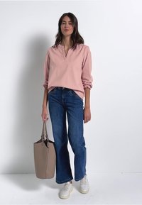 Helles rosa langärmeliges Oberteil mit V-Ausschnitt, dunkelblaue weit geschnittene Jeans, weiße Sneaker und eine beigefarbene Tragetasche. Glatte Textur mit lässiger Passform.