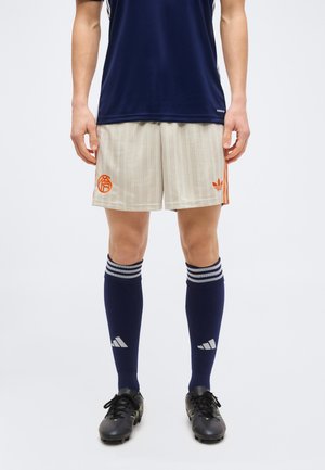 Άνδρας όρθιος που φοράει ναυτικό μπλε αθλητικές κάλτσες, μπεζ ριγέ σορτς με πορτοκαλί λογότυπα και μαύρα παπούτσια ποδοσφαίρου, μπροστά σε απλό φόντο.