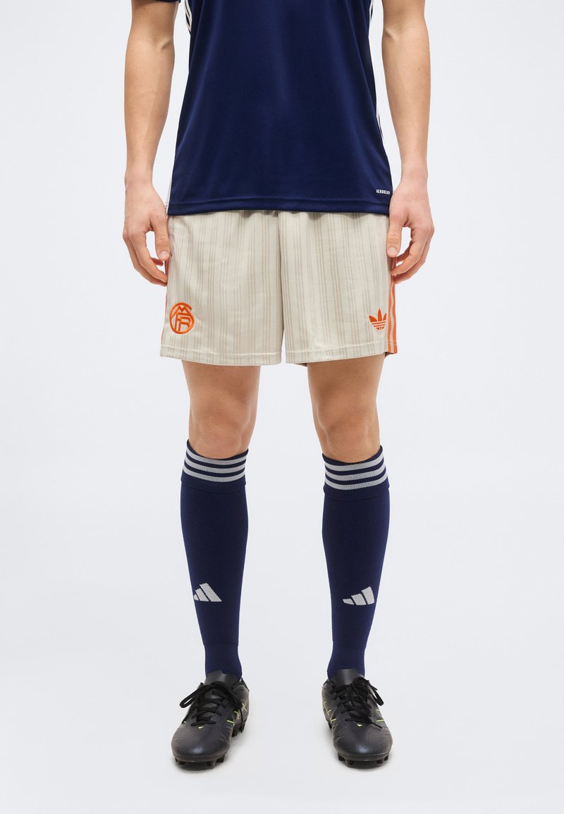 Hombre de pie que lleva calcetines deportivos azul marino, pantalones cortos beige a rayas con logotipos naranjas y tacos negros sobre un fondo liso.