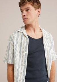 Joven con pelo rizado corto mira a la derecha, vistiendo una camisa de manga corta blanca a rayas sobre una camiseta de tirantes azul marino, frente a un fondo neutro.