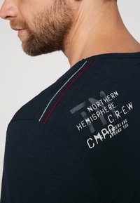 Camiseta azul marino con texto y diseño serigrafiado. Con costuras en rojo y gris a lo largo de la costura del hombro. La textura parece suave.