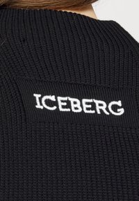 Černý pletený svetr s ribovanou strukturou a obdélníkovou bílou nášivkou se slovem "ICEBERG" vyšitým tučnými písmeny.