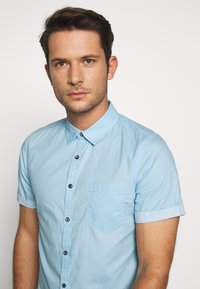 Hellblaues Kurzarmhemd mit einem dezenten Muster, Button-Down-Kragen, kleiner Brusttasche und schwarzen Knöpfen zum Schließen.