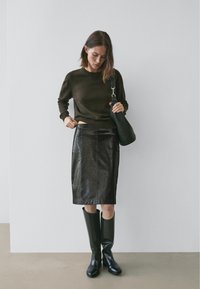 Jupe crayon en cuir noir avec une légère brillance, associée à un pull vert foncé et des bottes noires montantes. Tient un sac noir texturé.