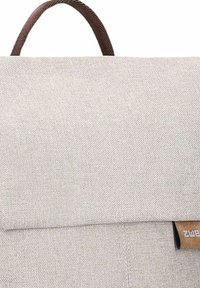 Mochila de tela gris claro con un asa marrón, tapa plegable y detalles de costura. Presenta una pequeña etiqueta marrón en el lado.