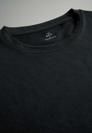 Čierne bavlnené tričko s okrúhlym výstrihom, s hladkou textúrou a minimalistickou etiketou s nápismi "THE SET" a "MADE IN BANJA LUKA."