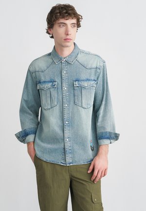 Jasnoniebieska koszula dżinsowa z ostrym kołnierzykiem, dwoma kieszeniami na piersi i zapięciem na guziki. Posiada podwinięte rękawy oraz małą ozdobną naszywkę.