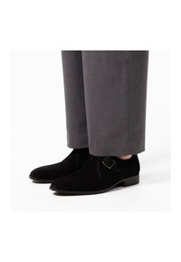 Black suede shoes with a rounded toe and a side buckle detail, paired with gray trousers, showcasing a smooth texture and simple design.