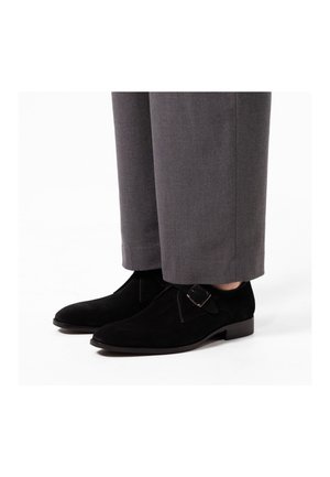Schwarze Wildlederschuhe mit einer abgerundeten Zehenform und einem seitlichen Schnallendetail, kombiniert mit grauen Hosen, die eine glatte Textur und ein schlichtes Design zeigen.