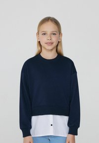 Marineblaues, verkürztes Sweatshirt mit langen Ärmeln, das über einem weißen Shirt getragen wird. Weicher Stoff, schlichtes Design, mit rundem Ausschnitt und Knopfdetail.