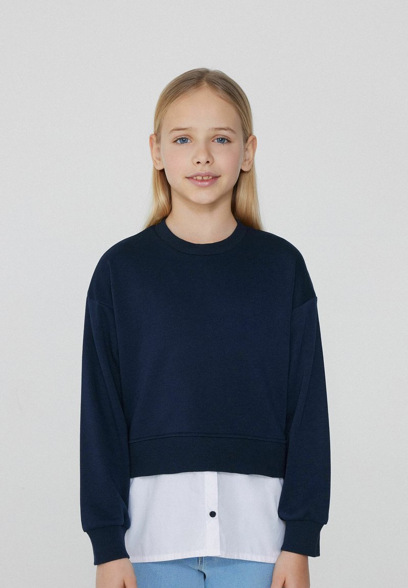 Marineblaues, verkürztes Sweatshirt mit langen Ärmeln, das über einem weißen Shirt getragen wird. Weicher Stoff, schlichtes Design, mit rundem Ausschnitt und Knopfdetail.