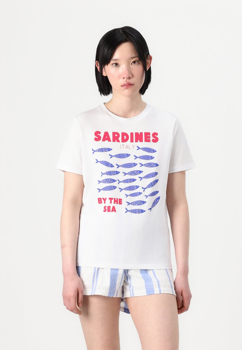 T-shirt de algodão branco com uma ilustração de peixes azuis e o texto “SARDINHAS (ITÁLIA) À BEIRA-MAR” em vermelho. Manga curta, ajuste solto.