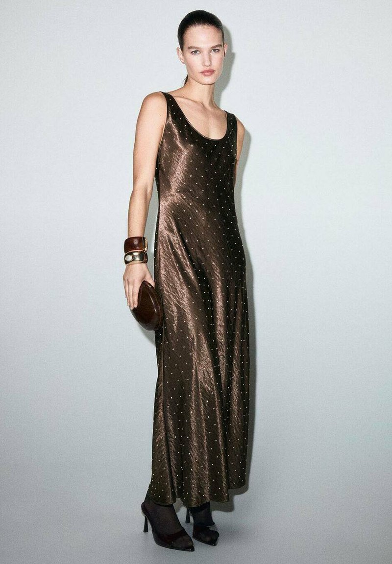 Vestido largo de satén marrón adornado con pequeños puntos metálicos, escote redondeado y una silueta fluida. Combinado con un bolso marrón y tacones altos.