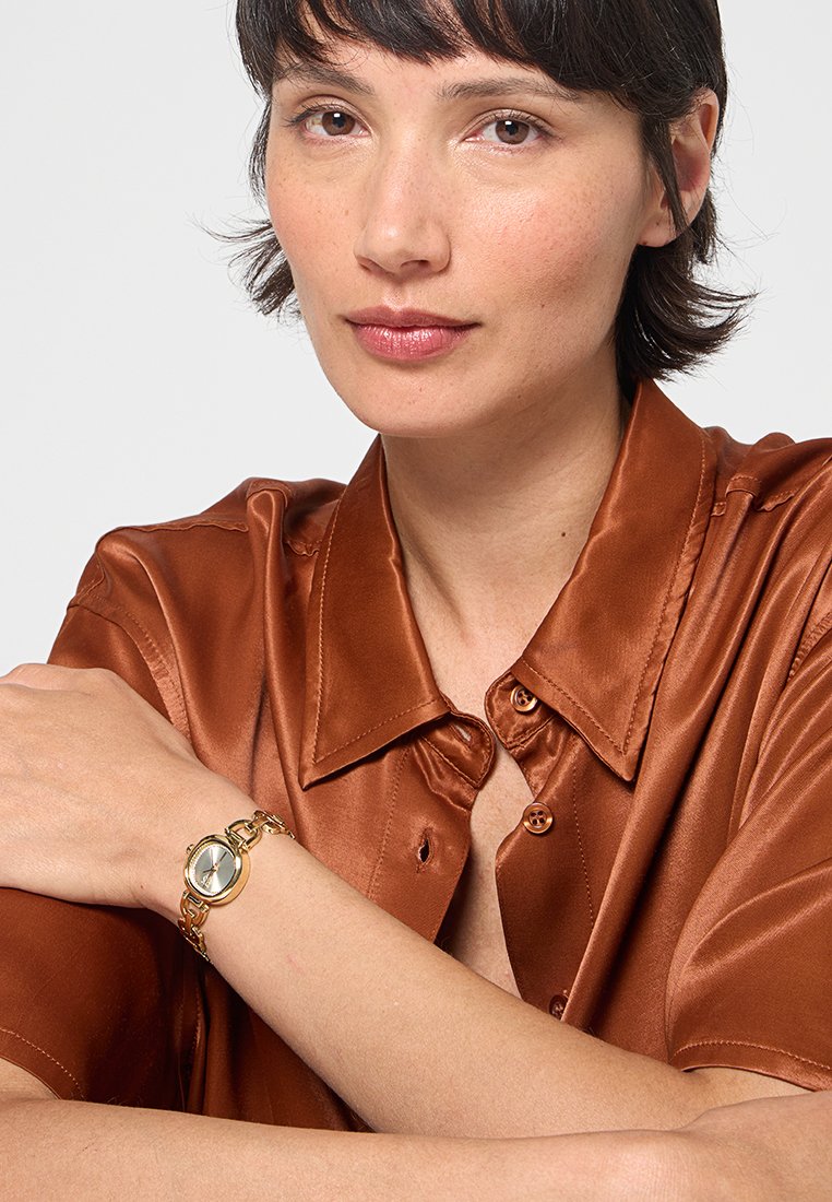 Reloj de pulsera dorado con cara redonda, correa de eslabones y una superficie reflectante, llevado en una muñeca sobre una camisa de satén marrón.