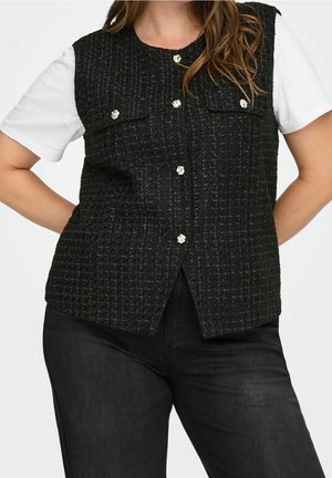 Mujer con chaleco negro texturizado con botones sobre camisa blanca de manga corta y pantalones negros, de pie con las manos en las caderas.